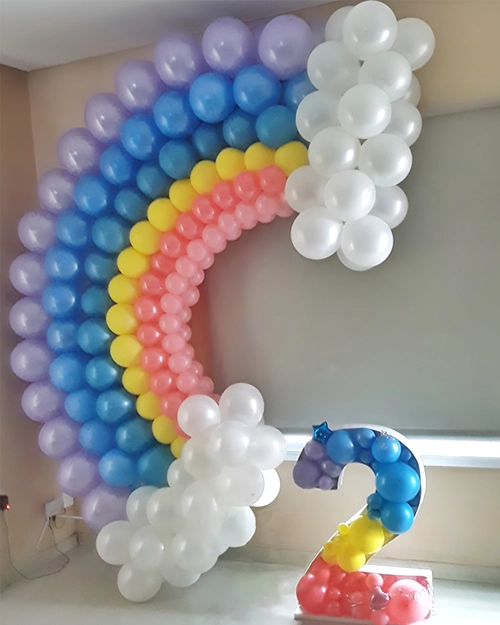 Semiarco con forma arcoiris de globos color gris, celeste, azul, amarillo, rosa y rosa claro. Nubes de globos blancas en los extremos. Número dos relleno de globos.