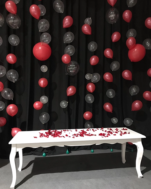 Cortina de globos rojos y translúcidos delante de una cortina negra. Por delante, una mesa vintage blanca con pétalos de rosa