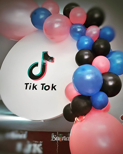 Círculo blanco con logo de TikTok adornado con globos Celestes, rosas y negros.