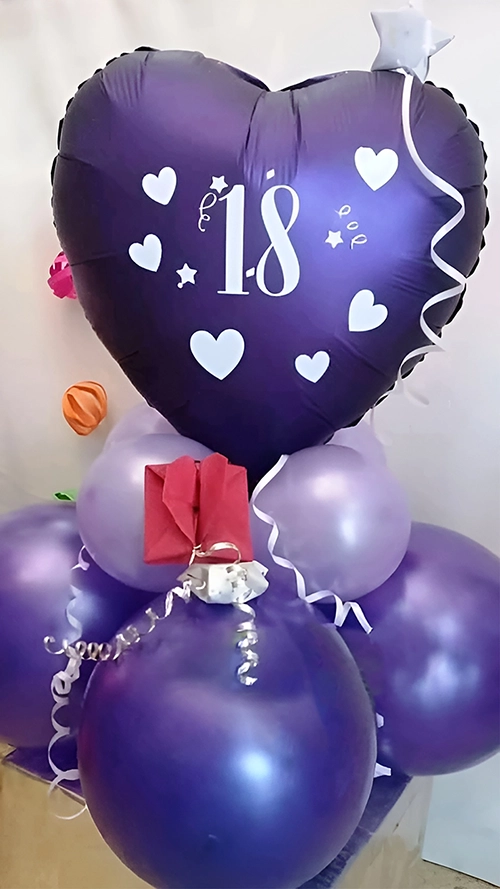 Bouquet festejo de 18 años. Globo corazón violeta oscuro estampado con corazones y el número 18 color blanco, sobre base de globos lilas y violetas. Cintas plateadas y sobre origami roja corazón decorando.