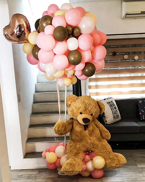 Globos tejidos formando un gran globo, atado a un oso de peluche marrón sobre base de globos. Colores marrones y rosados.