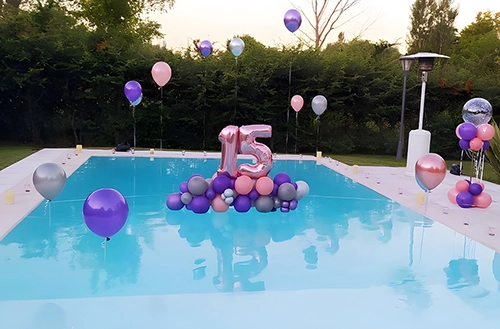 Decoración de cumpleaños de 15 en el jardín de una casa. Sobre una pileta, globos número 15 plateado sobre base globos opacos plateados, rosas y violetas. Globos helio decorando el borde de la pileta.